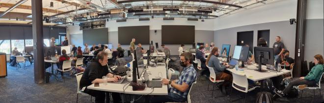 A panoramic shot of a room of people, some sitting at desks, some standing, some working on laptops, some in discussion.