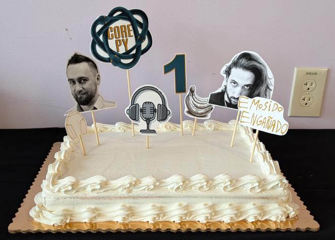 A big white cake, with decorations of Łukasz and Pablo’s faces, along with the Core.py logo, a big digit 1, headphones, microphone, bananas, and “emosido engañado” graffiti.