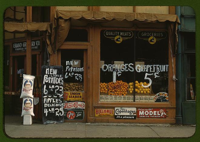 1942 store front of Grand Grocery Co. with many signs outside and lots of potatoes, oranges and grapefruit in the window. There are signs for Union Leader, Pepsi Cola, 7up, Beech Nut, Chew Copenhagen ('It's a pleasure') and Model Smoking Tobacco ('Did you say 10c?').
