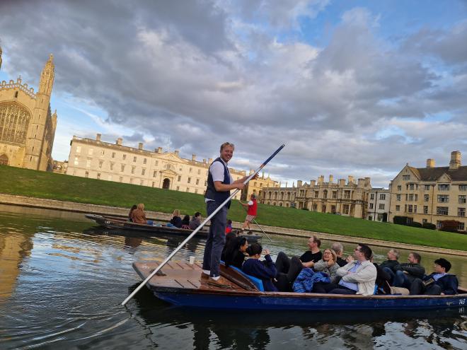 People sitting in a small boat, with a man standing at the back with a long pole in the water, in front of grand Cambridge college