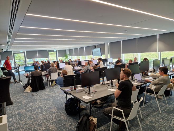 A room of people at desks with monitors. Steve has a mic and is asking a question to Brett who&rsquo;s standing at and presenting.